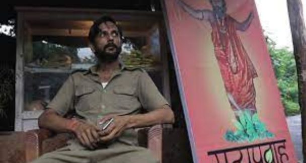 Garbage (2018) Antagonist Phaneeswar sitting next to a poster of his 'baba'